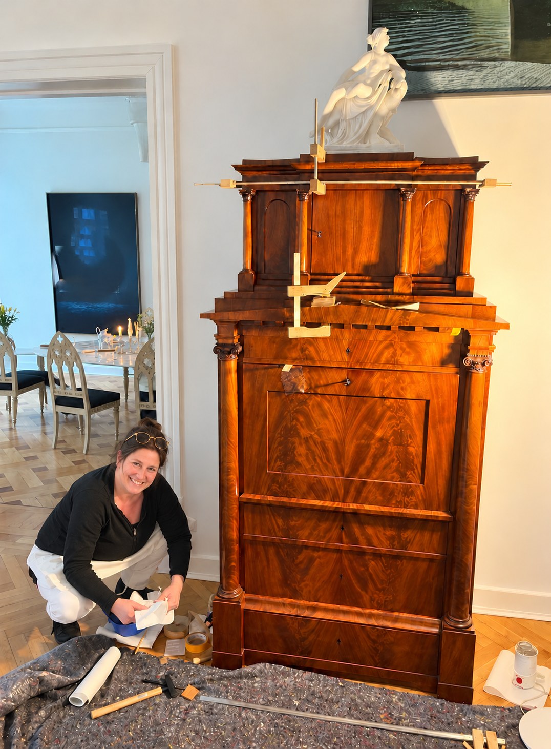 Hoher historischer Schreibschrank aus dunklem, stark gezeichnetem Holz mit Holzspanngurten im Aufsatz; davor kniet die Restauratorin mit weißem Tuch über einer kleinen blauen Schale, im Vordergrund eine Malerdecke mit Werkzeug